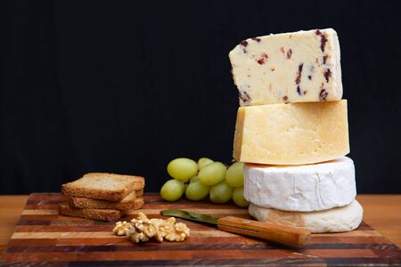 Stack of whole and cut cheeses and snacks, nuts, grapes, bread on chopping board against dark wall background. Closeup, side view, copy space. Cooking or gourmet conceptの写真素材