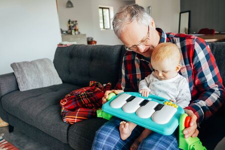 Happy grandfather playing with baby girl in living room. Senior man holding little child on lap with piano toy. Grandparents conceptの写真素材