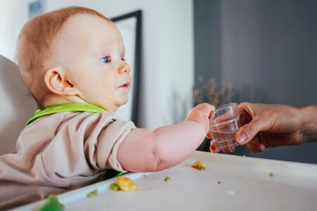 Mother giving glass of water to baby girl while feeding. Cute little child sitting on highchair and having breakfast. Led weaning conceptの写真素材