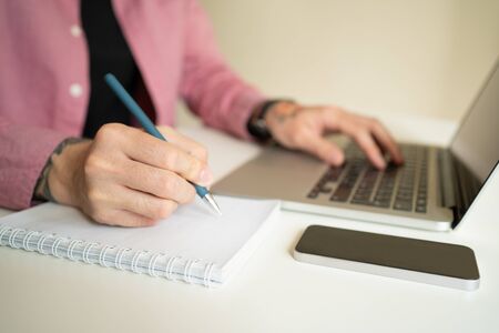 Unrecognizable young man in pink shirt writing with pen and typing on laptop. Guy with rose tattoo...