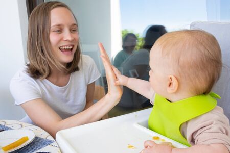 Happy mom and baby daughter giving high five to each other while having family lunch or dinner on balcony. First solid food or child care at home conceptの写真素材