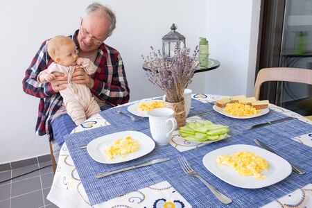 Positive grandpa sitting at dining table with food and holding baby granddaughter in arms. Family or babysitting conceptの写真素材