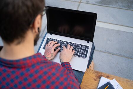 Bearded man sitting on wooden bench, typing on laptop and looking at screen. Hipster with rose tattoo on hand. Notebooks and pencil near him. Working outside conceptの写真素材