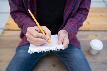 Unrecognizable man writing in notebook with pencil. Guy with tattoo on hand. Takeaway coffee...
