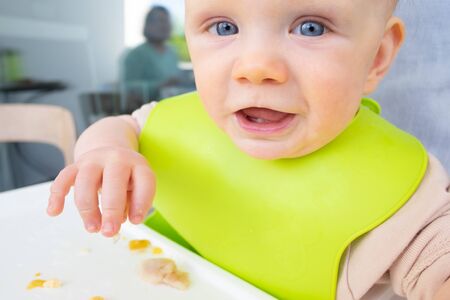 Closeup of happy baby wearing plastic bib, sitting in high chair and looking at camera. First solid food or child care at home conceptの写真素材