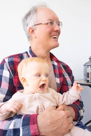 Happy laughing grandpa holding adorable baby granddaughter on his knees. Closeup, vertical shot. Family or babysitting conceptの写真素材