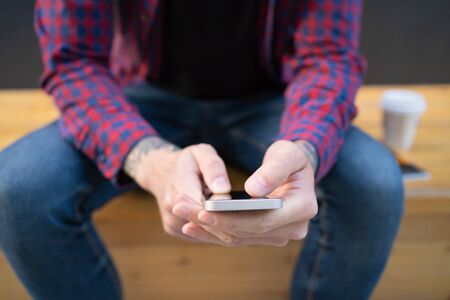 Unrecognizable man sitting on wooden bench with phone. Guy with tattoo on hand. Takeaway coffee...