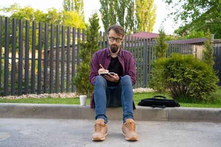Dark-haired man sitting on road curb and writing in note with pencil. Guy in eyeglasses and casual clothes. Grass on background. Working outside and freelance conceptの写真素材