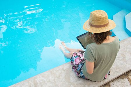 Female tourist in summer straw hat sitting at poolside, using tablet with pen, writing on screen....