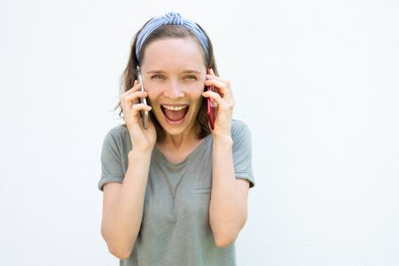 Excited laughing young woman shouting for joy while talking on two cellphones, looking at camera....