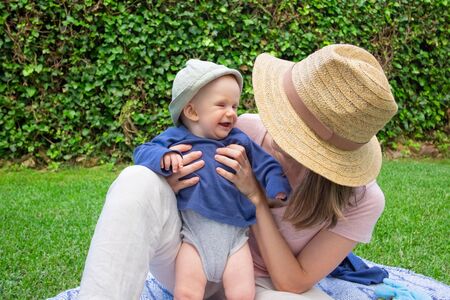 Young mom with face covered by hat holding daughter and talking with her in park. Cute red-haired...