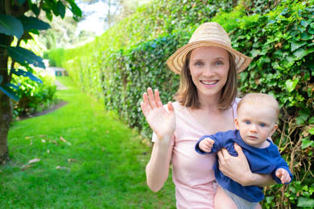 Beautiful mother in hat waving, holding newborn, smiling and looking at camera. Adorable baby on mom hands looking seriously. Summer family time, garden and sunny days conceptの写真素材