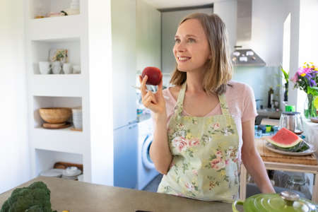Cheerful dreamy woman holding fruit, looking away and smiling while cooking in her kitchen. Copy...
