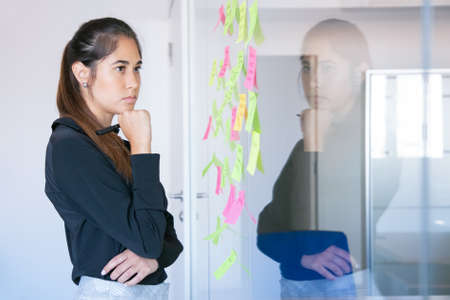 Thoughtful Latin businesswoman holding marker and reading notes on glass wall. Focused confident...