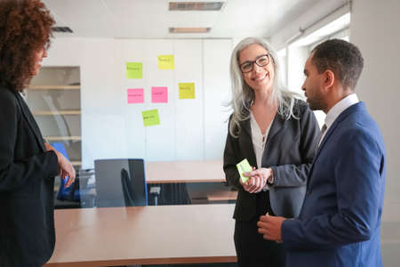 Content young businesspeople discussing marketing plan and making notes on stickers. Successful confident colleagues in suits meeting in office room. Teamwork, business and management conceptの写真素材