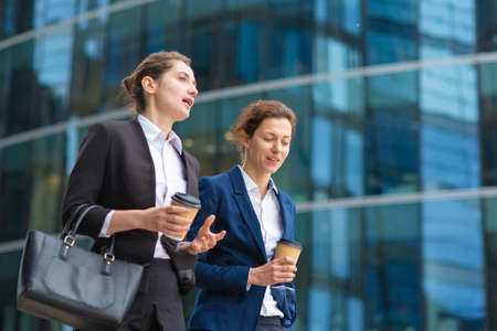 Young female professionals with takeaway coffee mugs wearing office suits, walking together past glass office building, talking, discussing project. Medium shot. Work break or friendship conceptの写真素材