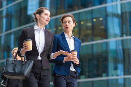 Young female employees with paper coffee cups wearing office suits, walking together past glass office building, talking, discussing project. Low angle. Work break or friendship conceptの写真素材