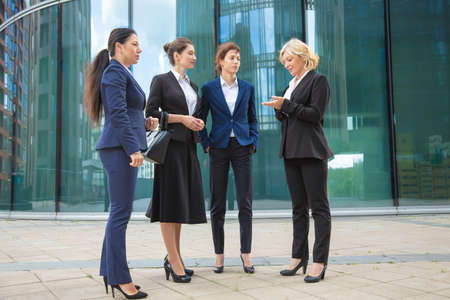 Successful female managers discussing project outdoors. Businesswomen wearing suits, standing together in city and talking. Full length, low angle. Cooperation and teamwork conceptの写真素材