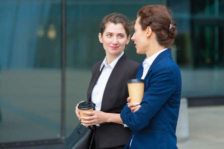 Positive office female friends with takeaway coffee cups walking together outdoors, talking, discussing project or chatting. Medium shot. Work break conceptの写真素材