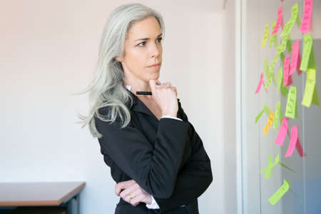 Thoughtful gray-haired businesswoman reading notes on glass wall and holding marker. Concentrated Caucasian female worker in suit thinking about idea for project. Brainstorming and business conceptの写真素材