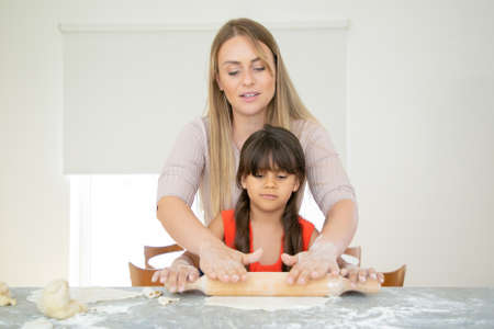 Focused girl and her mom rolling dough on kitchen table with flour powder. Mother teaching daughter...