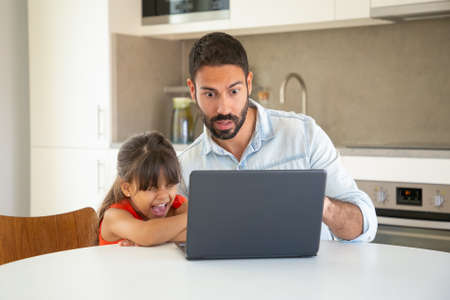 Excited girl and her surprised dad using laptop, sitting at table, watching horror or shocking...