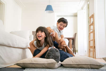 Happy dad and excited little kids having fun together, romping and playing on living room carpet, enjoying time at home. Family leisure time conceptの写真素材