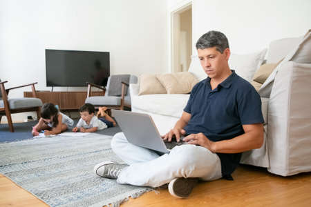 Focused dad working at home, sitting on floor and using computer while his little kids drawing in...