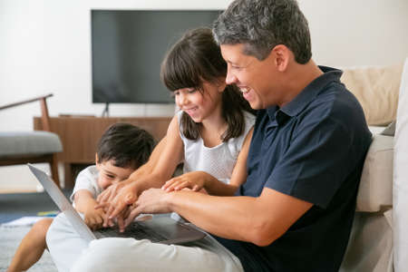 Joyful happy dad and two kids using laptop together, sitting on floor in apartment, pressing buttons...