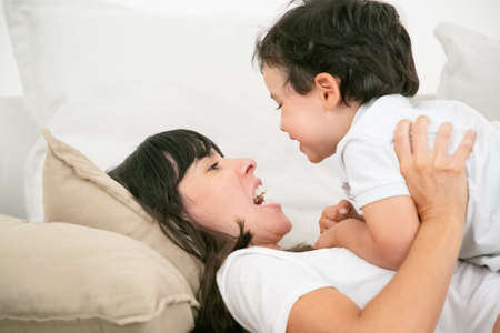 Young brunette mother lying on pillow and talking with cute son. Adorable little boy looking at...