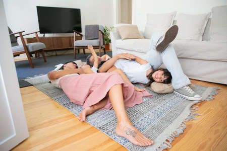 Happy family with two little children relaxing at home, lying on carpet on floor in living room, cuddling kids. Family fun time conceptの写真素材