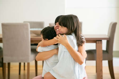 Happy mother hugging her lovely children with both hands. Pretty Caucasian mom sitting on floor with...