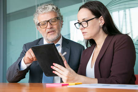 Female assistant showing report on tablet to male boss. Colleagues looking and pointing at gadget...