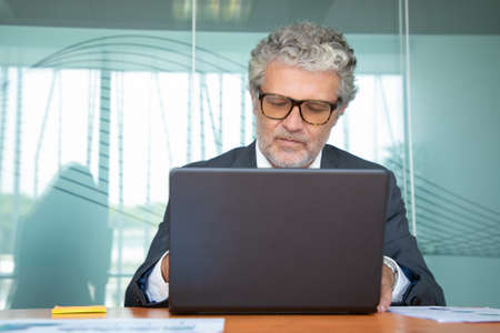Focused mature professional wearing suit and glasses, working at computer in office, using laptop at table. Medium shot, front view. Communication or wireless technology conceptの写真素材