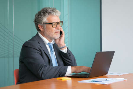 Serious mature executive in suit and glasses talking on mobile phone, working at computer in office,...