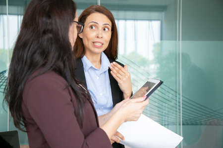 Professional businesswomen talking in conference room. Brunette middle-aged manager holding tablet....