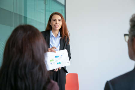 Successful beautiful employee standing and showing strategy plan. Experienced red-haired...
