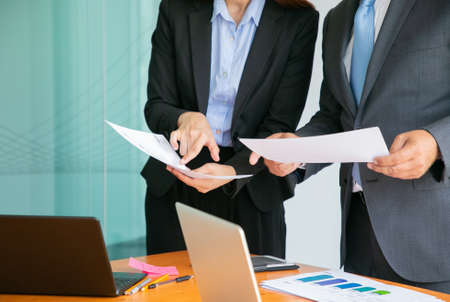 Unrecognizable entrepreneurs reading statistics and holding papers in hands. Successful professional partners standing near table with laptops. Business, management and partnership conceptの写真素材