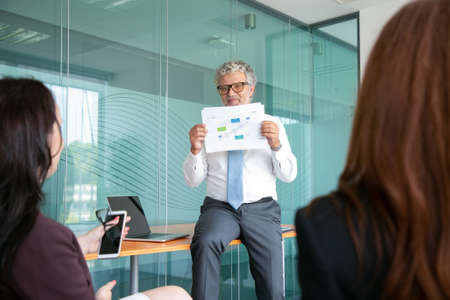 Professional bearded CEO sitting on table and showing graph. Experienced senior manager in eyeglasses holding paper, smiling and presenting project. Business and management conceptの写真素材