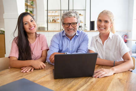 Happy successful business team sitting at table with open laptop, looking at camera, posing and...