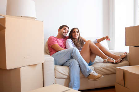 Happy Latin young couple relaxing on couch among cardboard packages in new apartment, looking away...