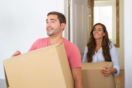 Joyful young Latin couple coming in their new apartment with carton boxes. Medium shot. Moving or...