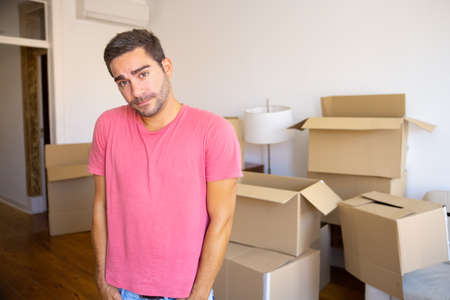 Pensive puzzled young man moving in new apartment, standing in front heap of opened carton boxes,...