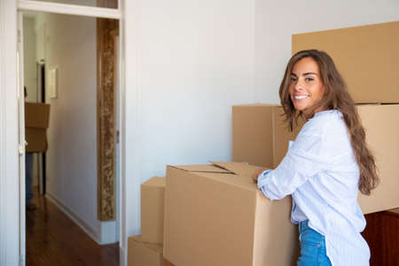 Cheerful beautiful young Hispanic woman unpacking stuff in her new apartment, standing near stacks...