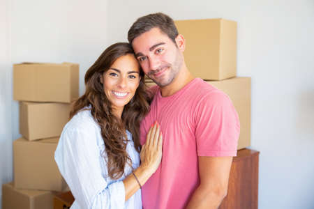Happy beautiful Hispanic couple standing among carton boxes in their new flat, hugging and looking...