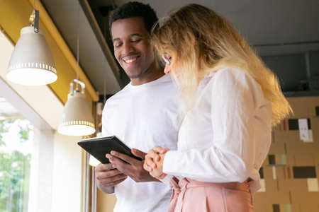 Happy African American designer presenting project on tablet screen and smiling. Long-haired blonde woman in glasses looking at device and listening to cheerful man. Communication and meeting conceptの写真素材
