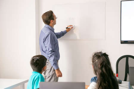 Back view of teacher writing on board and explaining lesson to pupils. Children sitting at table in classroom, looking at board and listening tutor. Childhood, study and education conceptの写真素材