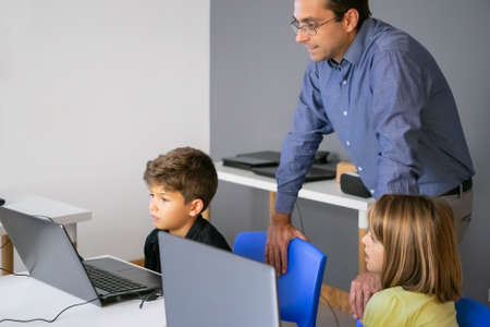 Lovely little pupil looking at laptop screen. Caucasian middle-aged teacher standing near him and classmate sitting at table. They watching and listening boy. Knowledge and digital education conceptの写真素材