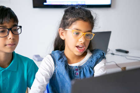 Mixed-race little girl sitting at table and reading task aloud. Concentrated in eyeglasses...