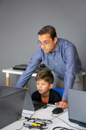 Content teacher checking task and standing behind pupil. Focused boy sitting at table in school,...